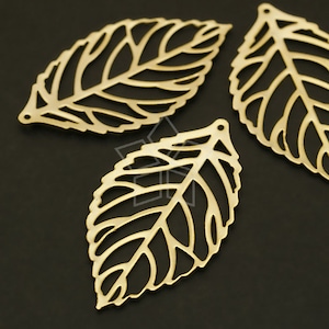 May include: Close-up of three gold-colored leaf-shaped charms. Each charm has a detailed, open-work design with veins and a small hole at the top for stringing. The leaves are arranged on a dark background.