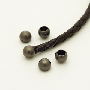 May include: Five dark gray metal beads with a hole through the center. The beads are arranged on a white surface. A brown leather cord is visible in the background.