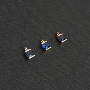 May include: Three square-shaped charms with blue gemstone accents. The charms are made of silver, gold, and rose gold.