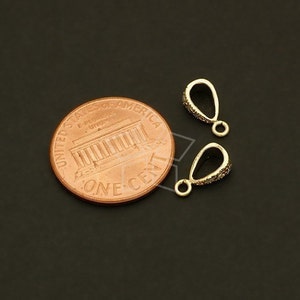 May include: Two gold-colored pendant bails with a teardrop shape, encrusted with small, sparkling stones. A US one-cent coin is next to the bails for size comparison. The bails have a small loop at the bottom for attaching to jewelry.