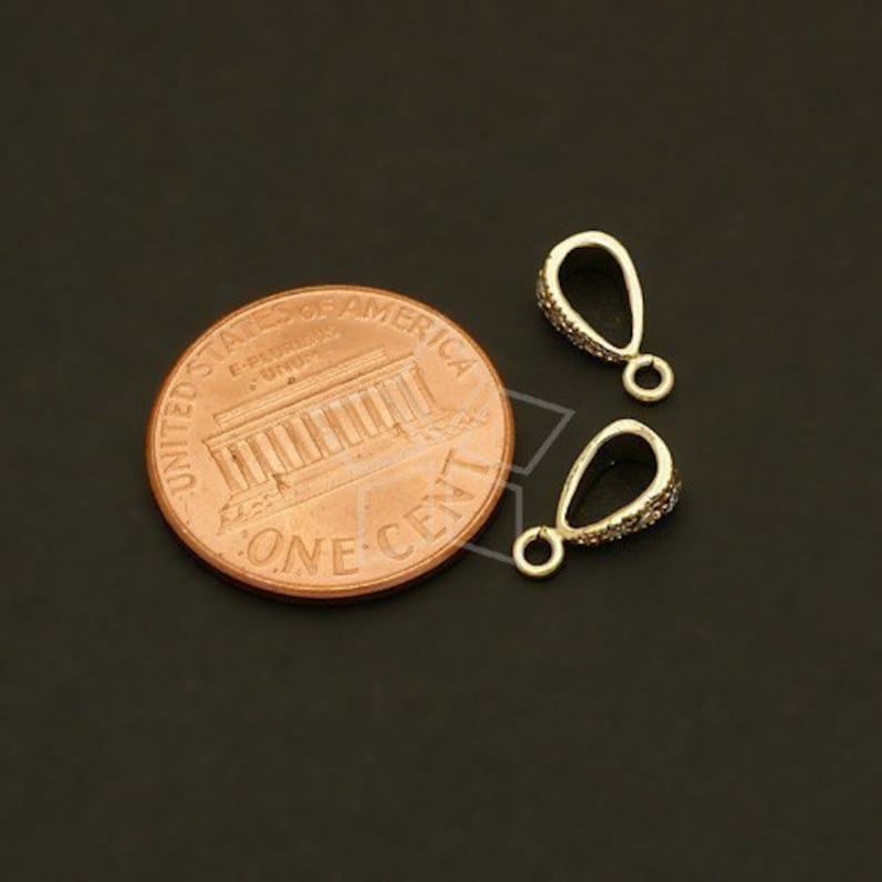 May include: Two gold-colored pendant bails with a teardrop shape, encrusted with small, sparkling stones. A US one-cent coin is next to the bails for size comparison. The bails have a small loop at the bottom for attaching to jewelry.