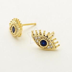 Könnte beinhalten: Goldfarbene Ohrstecker mit einem Augendesign. Jeder Ohrring hat eine blaue Mitte, umgeben von kleinen weißen Steinen und einem kronenartigen Oberteil. Die Ohrringe werden vor einem weißen Hintergrund präsentiert.