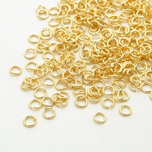 May include: A large pile of gold jump rings, scattered on a white surface. The jump rings are small and round, with a smooth finish.