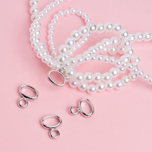 May include: Four silver oval-shaped jump rings with a smaller ring attached to each. The jump rings are on a pink surface with a strand of white pearl beads.