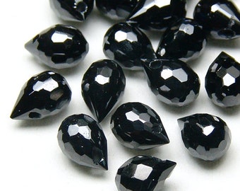 May include: A scattering of black, teardrop-shaped beads with a faceted surface. The beads have a glossy finish and are arranged against a white backdrop. The geometric cuts catch the light, creating a sparkling effect.