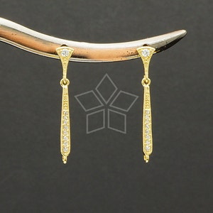 Può includere: Un paio di orecchini pendenti color oro. Ogni orecchino ha una parte superiore triangolare con una singola pietra trasparente e un corpo lungo e sottile a forma di goccia adornato con piccole pietre trasparenti. Gli orecchini sono esposti su uno sfondo scuro.