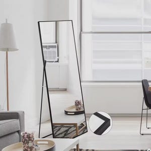 Op de afbeelding: Een zwarte, staande spiegel met een frame. De spiegel leunt tegen een witte muur. De spiegel reflecteert een kamer met een raam en een wit plafond.