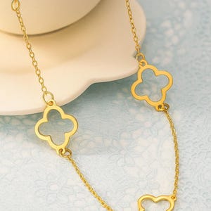 Peut inclure: Un collier doré avec une fine chaîne et des pendentifs en forme de trèfle à quatre feuilles ajourés. Le bijou est présenté sur un fond bleu clair à motifs, mettant en valeur le design et la couleur du collier.