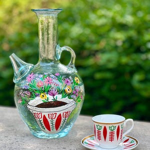 Puede incluir: Una jarra de vidrio transparente con un diseño floral y un patrón de tulipán rojo y blanco. Una taza de café de cerámica blanca con un patrón de tulipán rojo y blanco y un borde dorado se asienta sobre un platillo a juego.