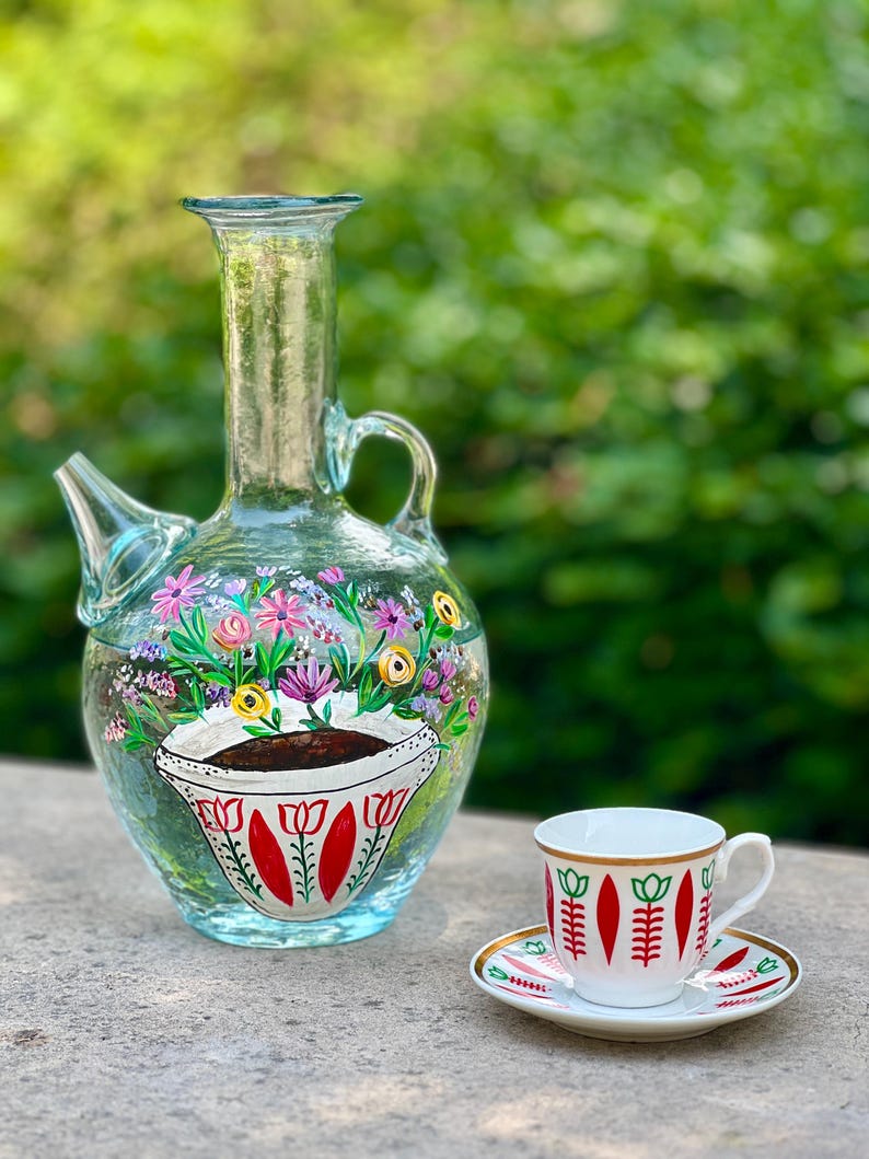May include: A clear glass pitcher with a floral design and a red and white tulip pattern. A white ceramic coffee cup with a red and white tulip pattern and a gold rim sits on a matching saucer.