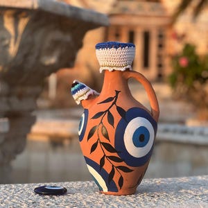 Può includere: Una brocca in ceramica color terracotta con un design a occhio blu e bianco e una parte superiore blu e bianca all'uncinetto. La brocca ha un manico e un beccuccio con una copertura all'uncinetto. Un piccolo pezzo di ceramica a forma di occhio abbinato è davanti.