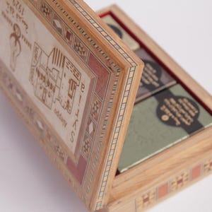 Puede incluir: Una caja de madera con un intrincado diseño de marquetería en la tapa y los lados. La caja está abierta, revelando cuatro pequeñas barras de jabón en el interior. La tapa presenta un diseño de un edificio con texto en un idioma extranjero.