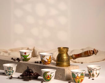May include: Six white ceramic coffee cups with colorful hand-painted designs, including a hamsa hand, a mosque, and a crescent moon. A traditional Turkish coffee pot with a wooden handle sits in the center of the image. The cups and pot are arranged on a gray concrete surface with coffee beans scattered around.