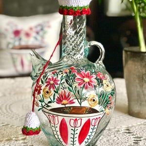 May include: A clear glass bottle with a crocheted white lid, painted with red and yellow flowers and a white cup with red accents. The bottle has a textured surface and a handle.