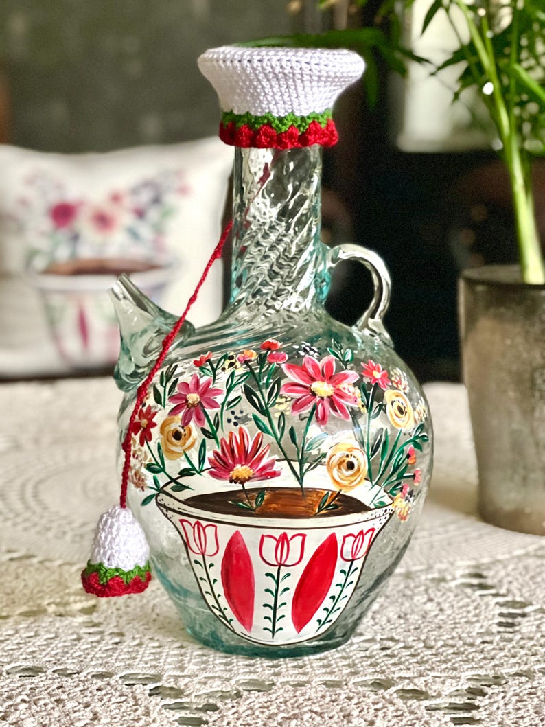 May include: A clear glass bottle with a crocheted white lid, painted with red and yellow flowers and a white cup with red accents. The bottle has a textured surface and a handle.