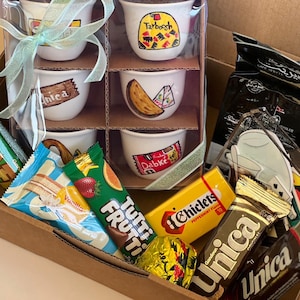 May include: A gift box featuring six small white coffee cups with decorative designs, a coffee package, and assorted snacks. The cups are in a cardboard holder, tied with a light green ribbon. Several individually wrapped sweets and wafers are also included.