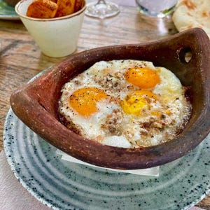 Può includere: Primo piano di una ciotola di terracotta piena di tre uova fritte, condite con pepe nero. La ciotola è su un piatto con un motivo grigio e bianco.