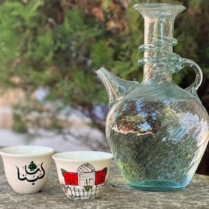 Op de afbeelding: Twee witte keramische koffiekopjes met traditionele Libanese ontwerpen. Een kopje heeft een groene cederboom en het Arabische woord "Libanon". Het andere kopje heeft een rood gebouw met een raam en een balkon. Achter de kopjes staat een heldere glazen kan met een getextureerd oppervlak.