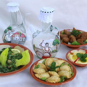 Könnte beinhalten: Ein Tischarrangement mit nahöstlichen Gerichten: Tabbouleh, Kibbeh, Hummus und Gebäck. Dazu zwei dekorative Glaskrüge mit bemalten Szenen und gehäkelten Deckeln. Die Speisen sind auf terrakottafarbenen Tellern angerichtet.