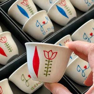 Op de afbeelding: Een collectie witte keramische kopjes met handgeschilderde ontwerpen. Sommige kopjes tonen een rode tulp met een groene steel en een rode ruitvorm, terwijl andere blauwe cursieve initialen met een klein rood hartje en de datum 05.29.20 bevatten.