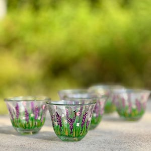 Könnte beinhalten: Ein Set aus klaren Glasbechern mit handgemalten Motiven von violetten Lavendelstängeln, grünem Gras und kleinen weißen Blüten. Jeder Becher verfügt über einen ausgestellten Rand und einen sich verjüngenden Boden.