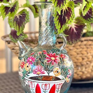 May include: A clear glass vase with a twisted neck and a floral design painted on the front. The vase is decorated with pink, yellow, and white flowers, and a cup of coffee with red and green accents.
