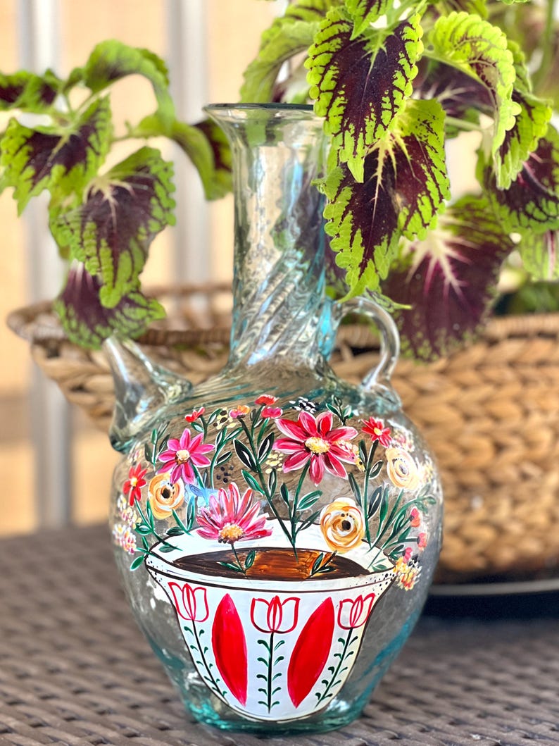 May include: A clear glass vase with a twisted neck and a floral design painted on the front. The vase is decorated with pink, yellow, and white flowers, and a cup of coffee with red and green accents.