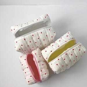 Könnte beinhalten: Vier weiße Kosmetiktaschen mit roten Herzmustern. Die Taschen sind geöffnet und zeigen weiße, gelbe und rosa Innenräume. Jede Tasche hat einen weißen Reißverschluss.