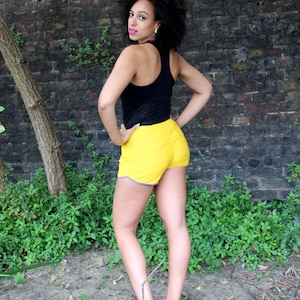 May include: A woman wearing a black tank top and yellow athletic shorts stands in front of a brick wall. She is wearing gray athletic shoes.