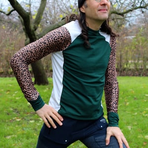 May include: A person wearing a green and white long-sleeved shirt with a leopard print on the sleeves. The shirt has a high neck and the person is wearing a black beanie and blue jeans.
