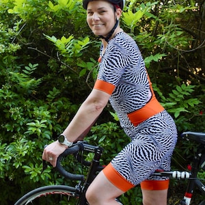 Puede incluir: Una persona con un traje de ciclismo blanco y negro con detalles naranjas y un casco negro está montando una bicicleta negra. La bicicleta tiene un sillín con estampado blanco y negro y un manillar negro.