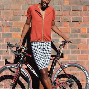 Może przedstawiać: Mężczyzna w czerwonej koszulce kolarskiej, czarno-białych szortach w kratę i czarno-białej czapce stoi obok czarnego roweru szosowego z czerwonymi akcentami. Rower ma czarno-białe siodełko i czarną kierownicę.