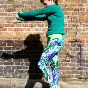 May include: A woman wearing a teal long-sleeved top and colourful leggings with a green, blue and purple pattern. She is standing in front of a brick wall and is stretching her arm.