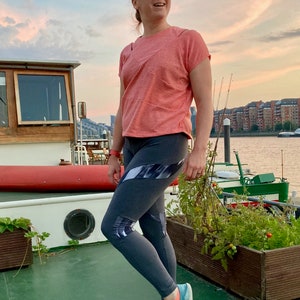 May include: A woman wearing a pink t-shirt and gray leggings with a black and white geometric pattern. She is standing on a boat deck with a green surface and a wooden planter with plants in the background.