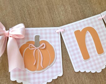 May include: A decorative banner featuring three pennants with a light pink and white gingham pattern. The first pennant displays an orange pumpkin with a pink ribbon, while the next two show the orange letters n and e, accented by pink bows.