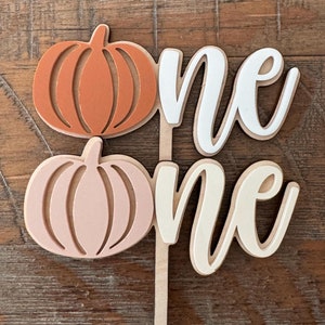 May include: A wooden cake topper with the word "one" written twice in white and cream. The topper is decorated with two pumpkins, one orange and one pink.