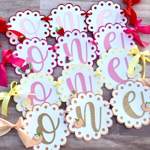 May include: A pink and yellow banner with the word "One" spelled out in individual letters. Each letter is on a separate white card with a scalloped edge. The letters are decorated with glitter, fruit, and flowers.