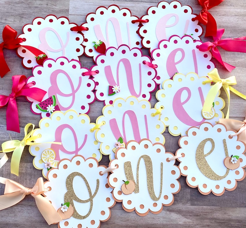 May include: A pink and yellow banner with the word "One" spelled out in individual letters. Each letter is on a separate white card with a scalloped edge. The letters are decorated with glitter, fruit, and flowers.