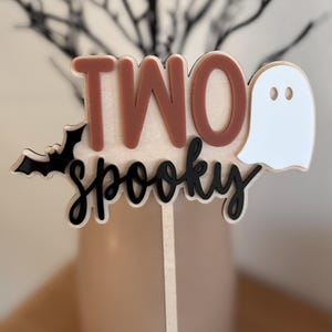 May include: A cake topper featuring the text TWO in terracotta and spooky in black script. The design includes a black bat on the left and a white ghost on the right, all mounted on a light wood backing with a thin wooden stick for insertion.