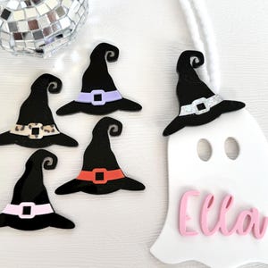 May include: Five black witch hats with different coloured accents, including leopard print, purple, orange, and white glitter. A white ghost with the name "Ella" written in pink letters.