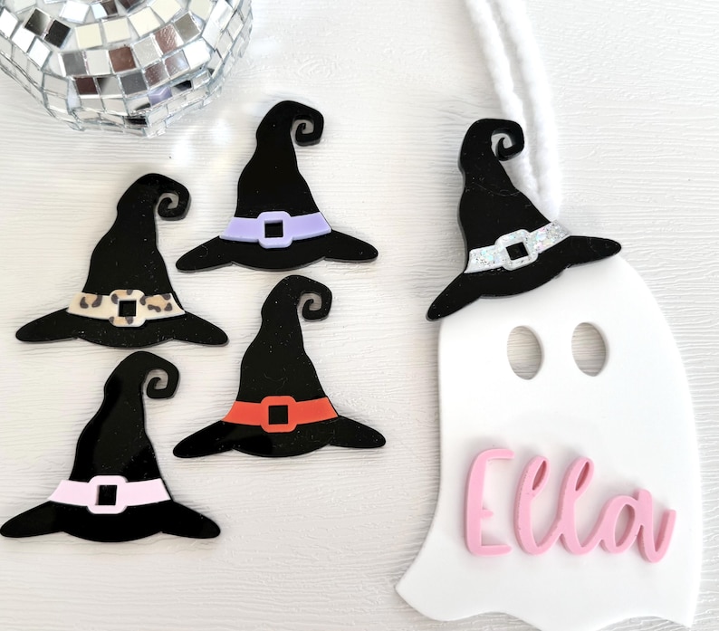 May include: Five black witch hats with different coloured accents, including leopard print, purple, orange, and white glitter. A white ghost with the name "Ella" written in pink letters.