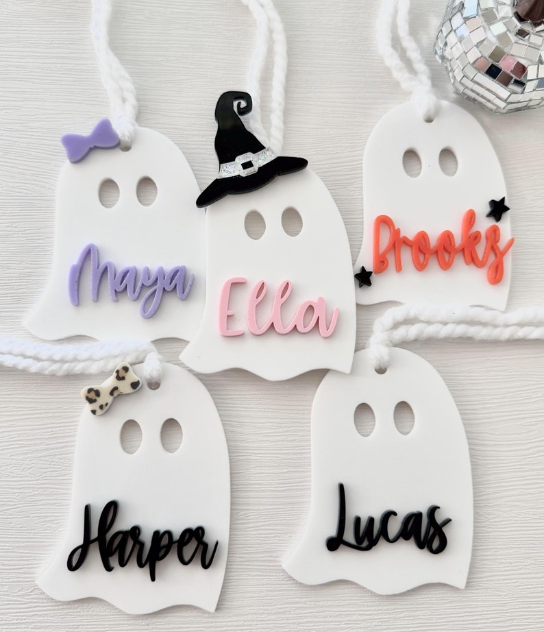 May include: Five white ghost-shaped ornaments with different names written on them. The names are Maya, Ella, Brooks, Harper, and Lucas. Some of the ghosts have additional decorations, such as a witch's hat, a bow, and a star.