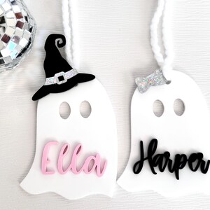 May include: Two white ghost ornaments with black and pink lettering. The ghost on the left has a black witch hat and the name "Ella" in pink. The ghost on the right has a silver glitter bow and the name "Harper" in black.