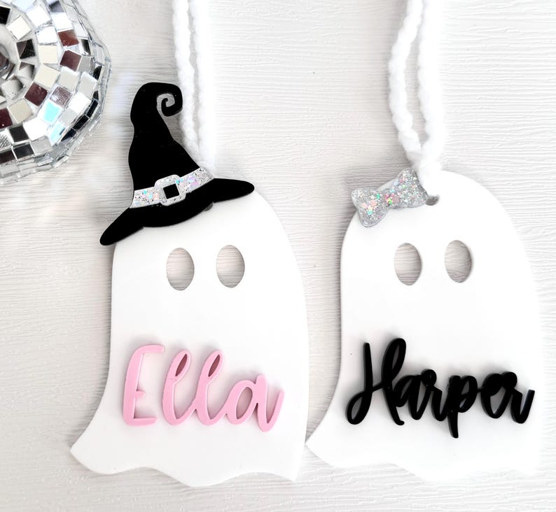 May include: Two white ghost ornaments with black and pink lettering. The ghost on the left has a black witch hat and the name "Ella" in pink. The ghost on the right has a silver glitter bow and the name "Harper" in black.