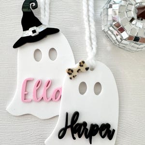 May include: Two white ghost-shaped ornaments with black and pink lettering. The top ghost has a black witch hat and the bottom ghost has a black and white bow. The names "Ella" and "Harper" are written on the ornaments.