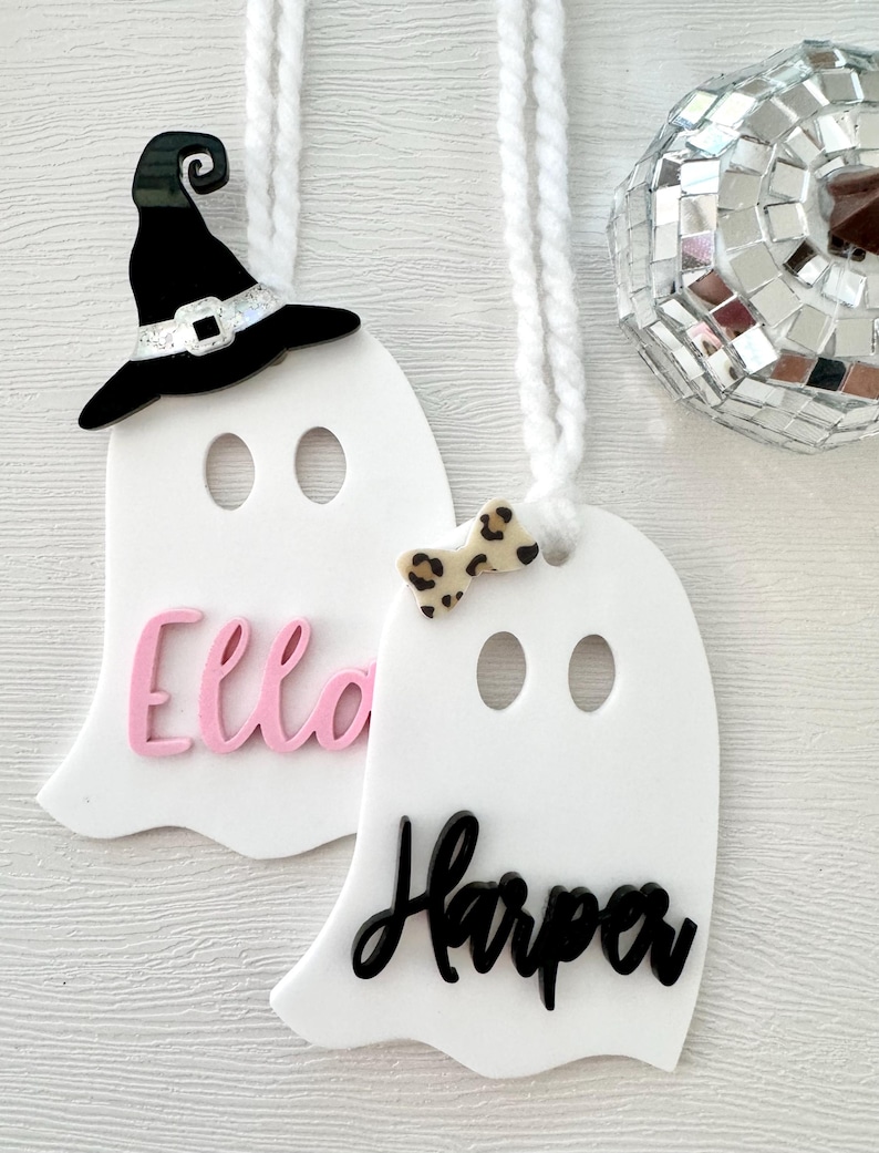 May include: Two white ghost-shaped ornaments with black and pink lettering. The top ghost has a black witch hat and the bottom ghost has a black and white bow. The names "Ella" and "Harper" are written on the ornaments.