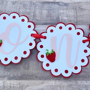 May include: A red and white banner with the word "one" in pink script. Each letter is on a separate circle with a red scalloped edge and red polka dots. Each circle also has a red strawberry with a green leaf and a white flower.