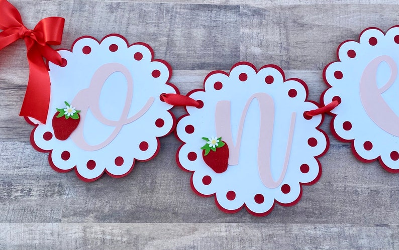 May include: A red and white banner with the word "one" in pink script. Each letter is on a separate circle with a red scalloped edge and red polka dots. Each circle also has a red strawberry with a green leaf and a white flower.