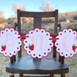 May include: A red and white banner with the word "one" in pink letters. Each letter is on a separate circle with a scalloped edge and red polka dots. There is a strawberry with a green leaf and white flower on each circle.