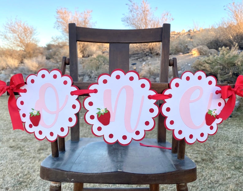 May include: A red and white banner with the word "one" in pink letters. Each letter is on a separate circle with a scalloped edge and red polka dots. There is a strawberry with a green leaf and white flower on each circle.